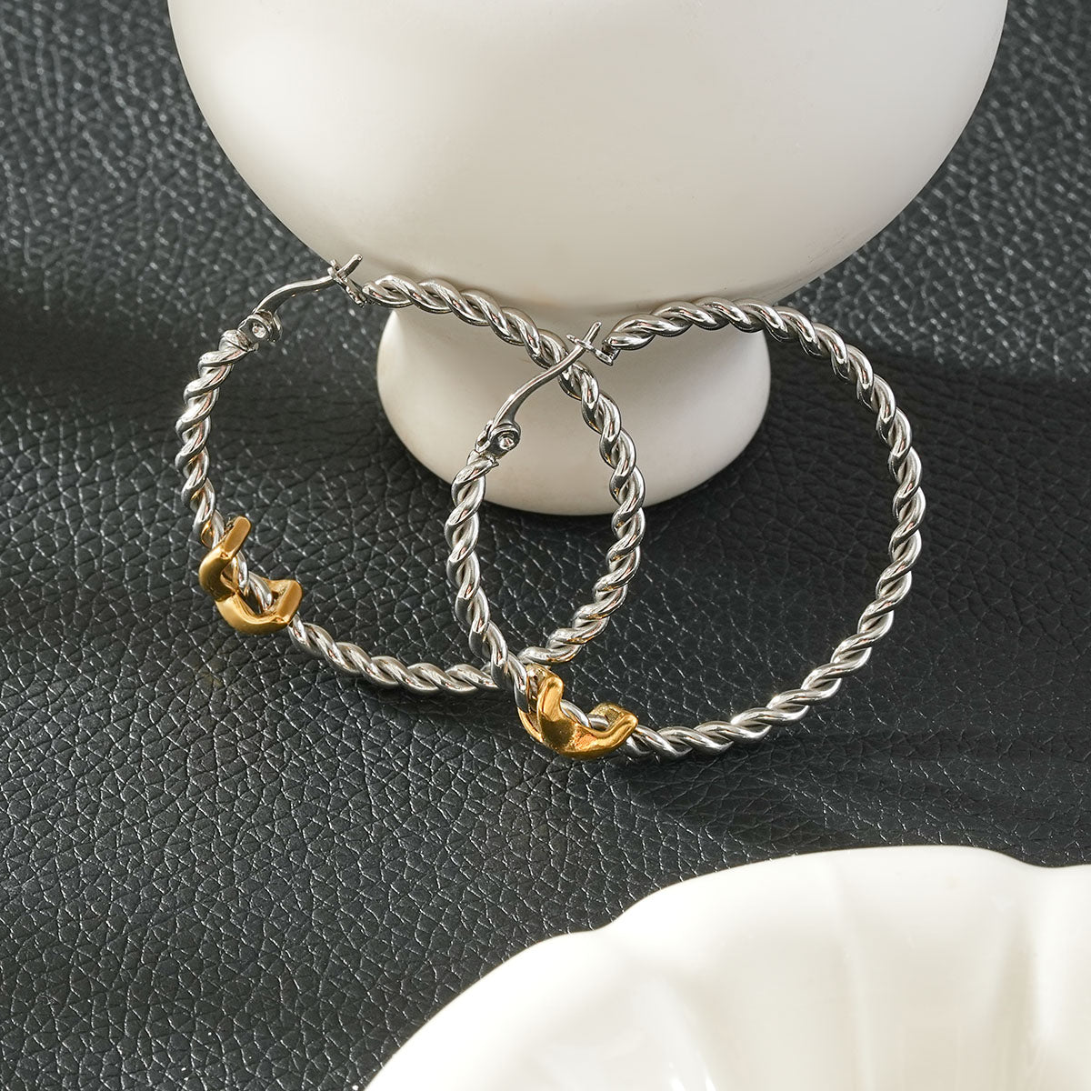 CIPRIANNA silver-tone rope twist hoop earrings with polished gold accents displayed on a textured black background.