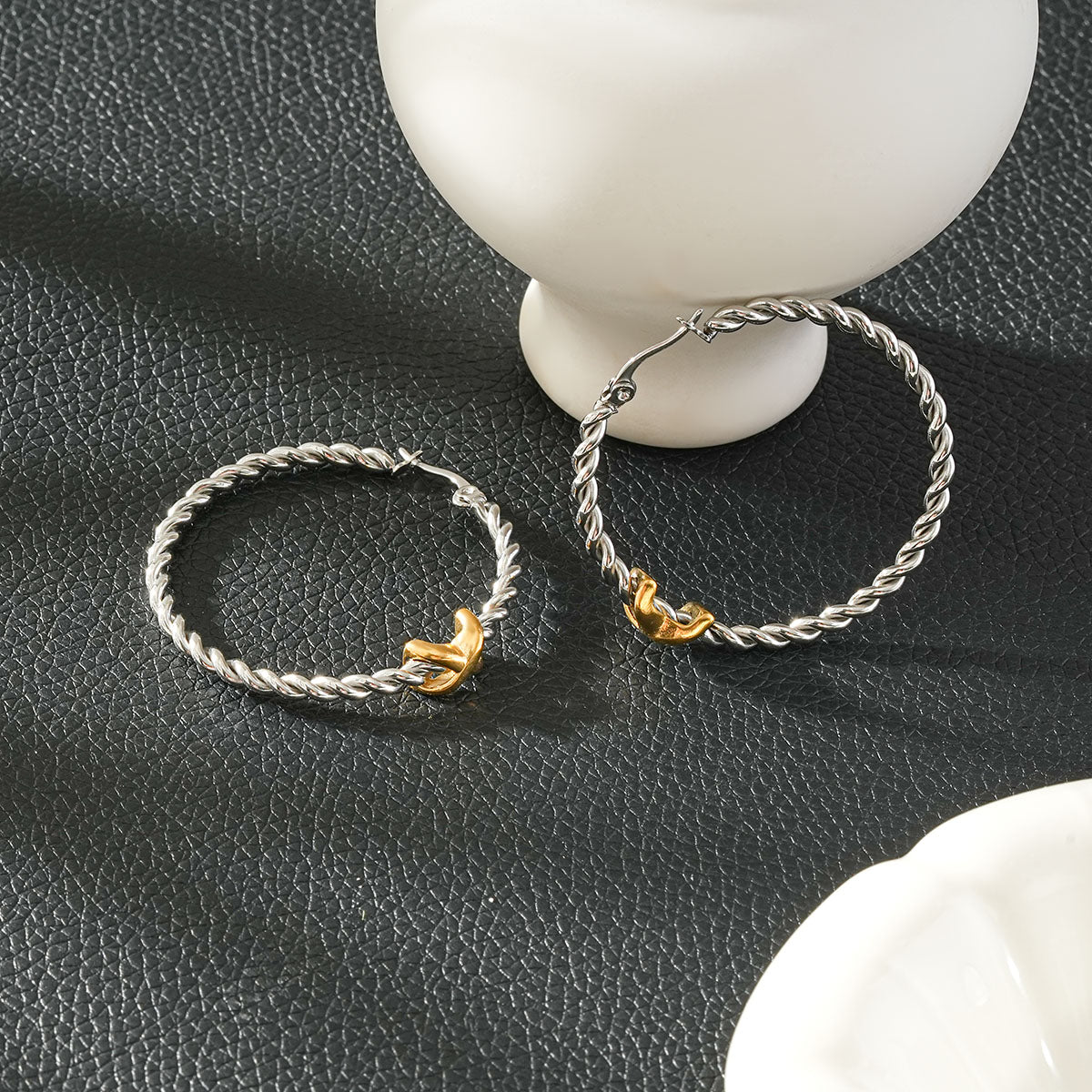 CIPRIANNA silver-tone rope twist hoop earrings with polished gold accents displayed on a textured black background.